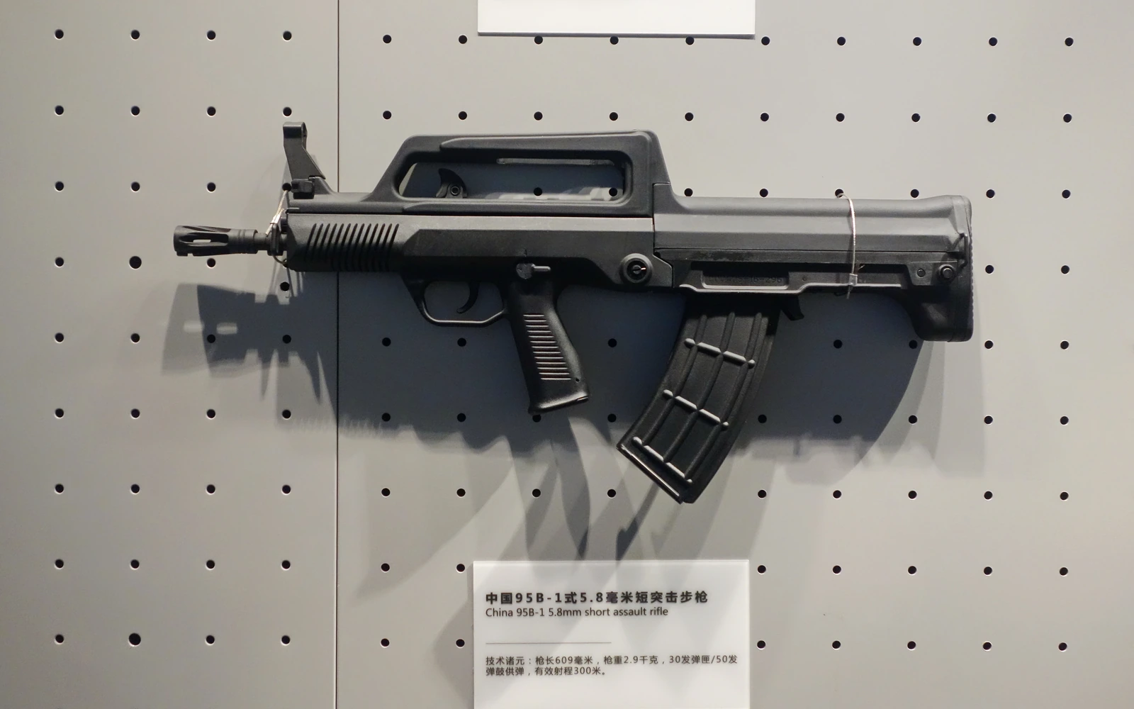 A short bullpup rifle mounted on a museum display wall, seen from the left, with a carrying-handle sight, a curved magazine behind the trigger and a Chinese and English label below