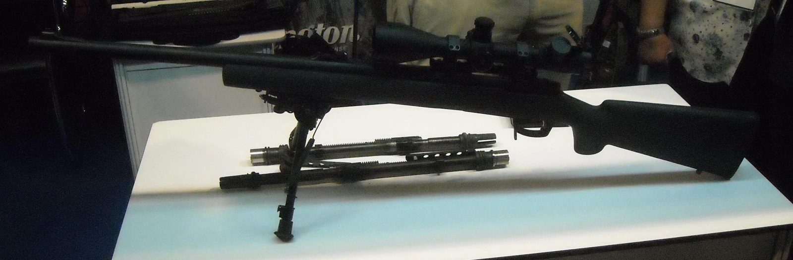 A bolt-action rifle with a wooden stock, a mounted telescopic sight and a bipod attached under the forend, resting on a table.