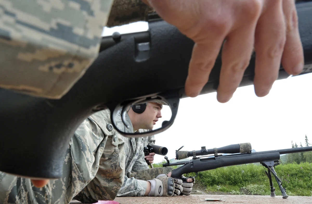 A serviceman in camouflage lies prone behind a scoped bolt-action rifle on a shooting range, sighting toward the camera.
