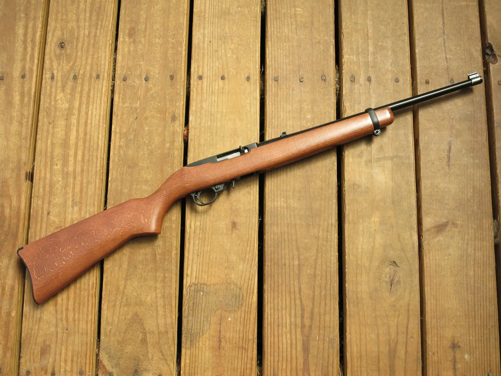 A Ruger 10/22 rifle lying on weathered wooden decking, seen from the left, with a plain hardwood stock, a barrel band and a rotary magazine sitting flush with the underside of the stock