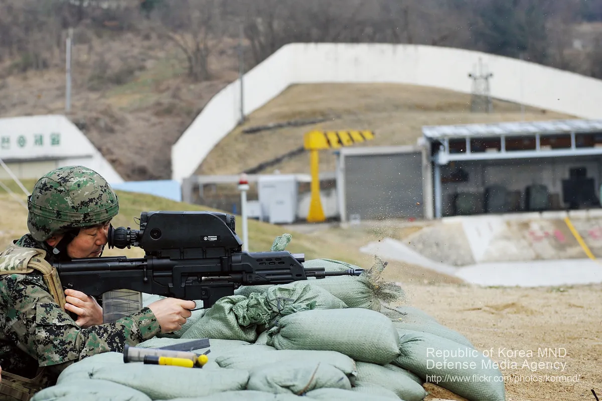 A soldier firing a K11 rifle from a prone position on an outdoor range, sandbags in front