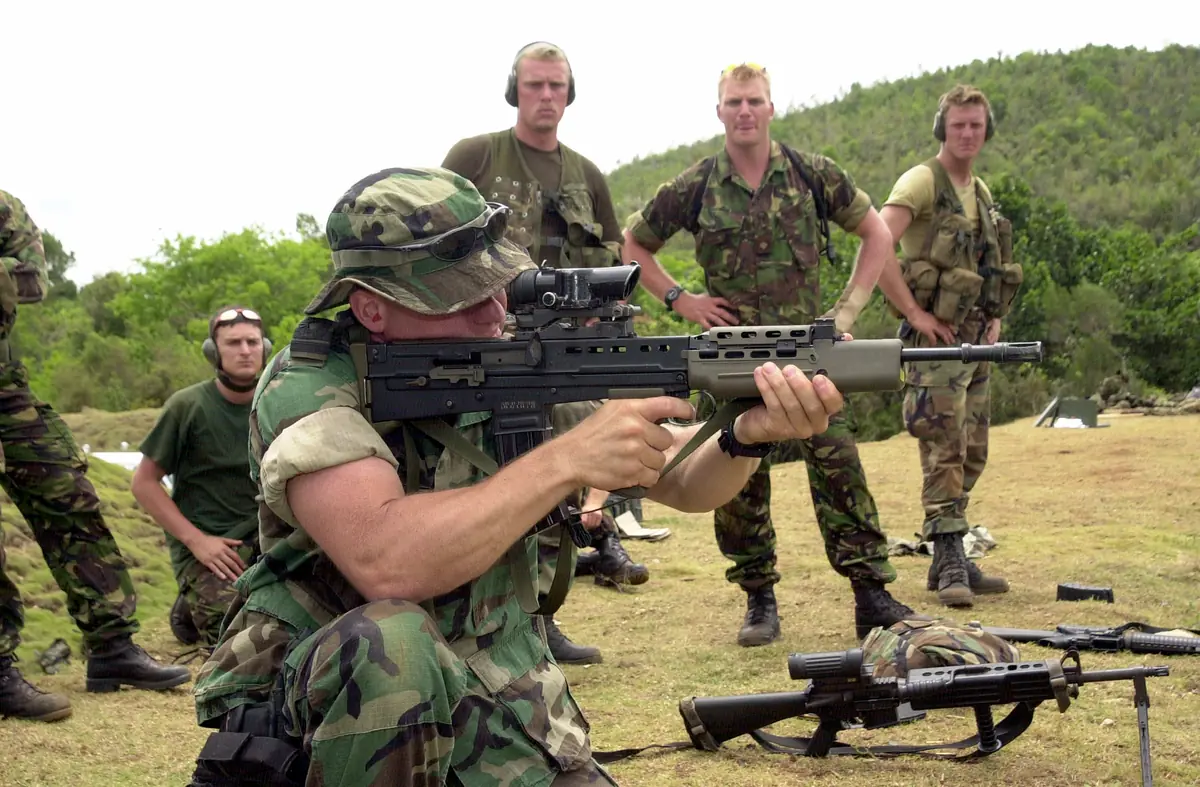 A soldier lying prone on gravel, aiming a black bullpup rifle downrange with an optical sight mounted above the barrel