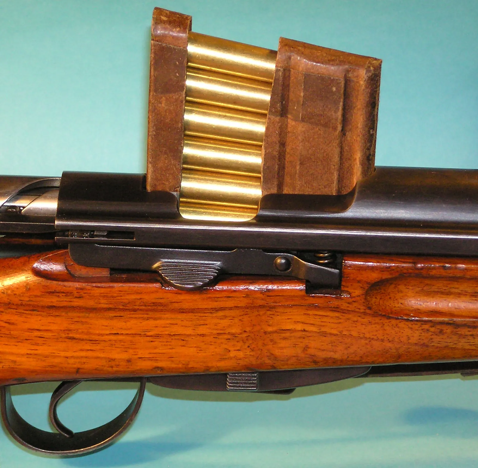 A close-up of the open breech of a K31 rifle on a pale blue background, with a six-round charger clip of brass cartridges held in a card sleeve inserted into the receiver above the magazine