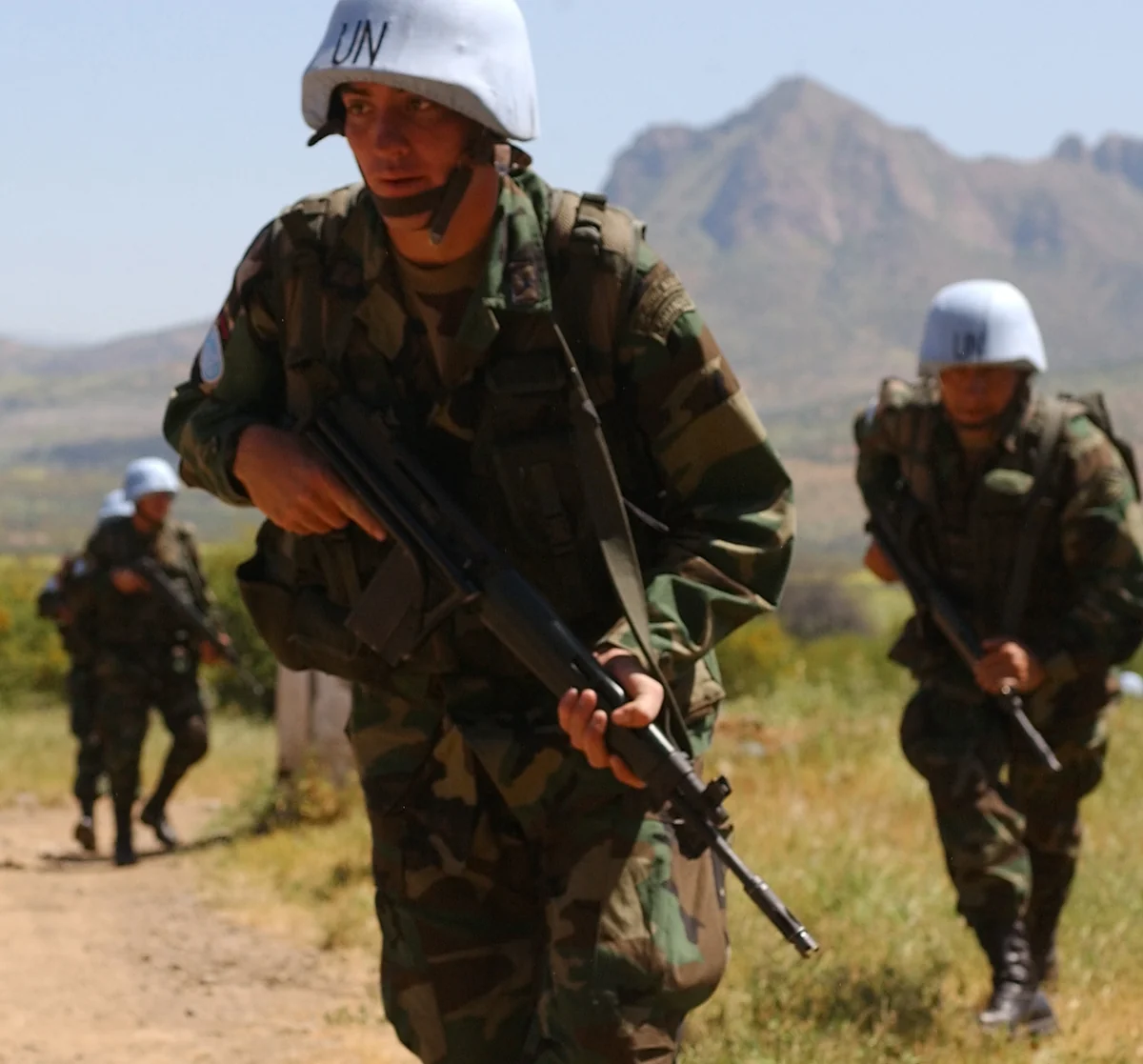 Soldiers in woodland camouflage and pale blue helmets marked UN advance across a dry field below mountains, the nearest carrying a black rifle at the ready.