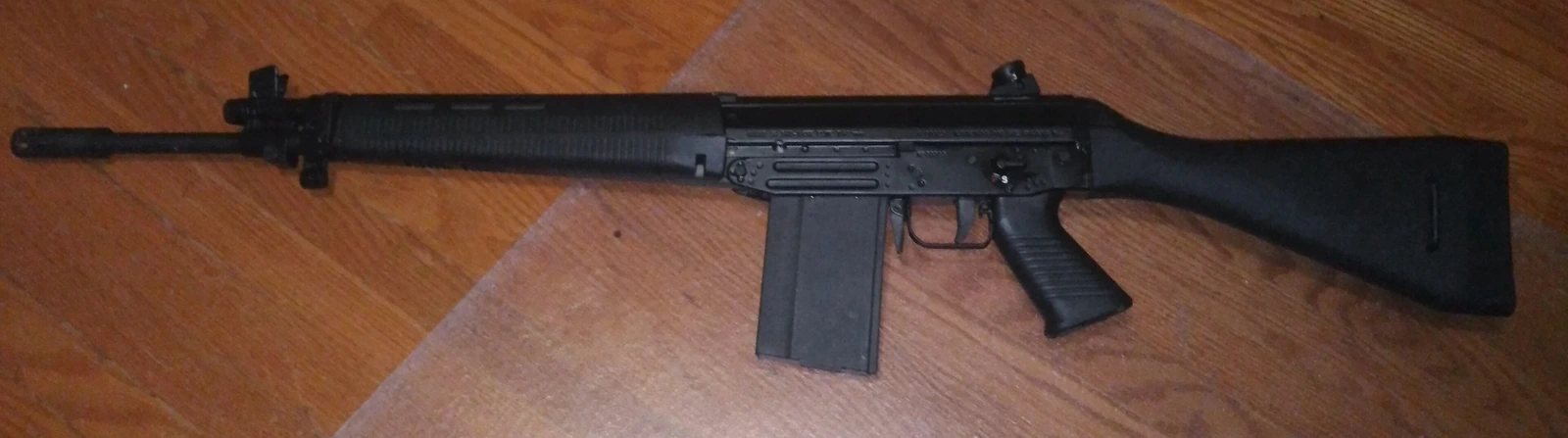 A black rifle with a fixed polymer stock, pistol grip, straight box magazine and a long flash suppressor, lying on its right side on a wooden floor.