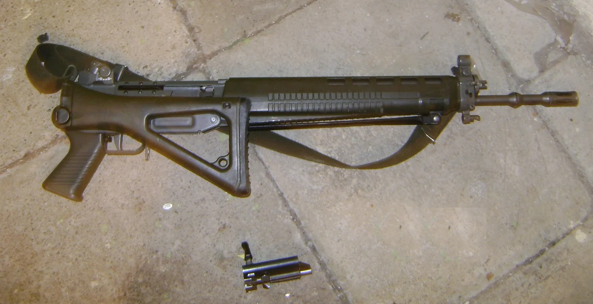 The rear of a SIG SG 550 rifle with its triangular wire stock folded flat against the side of the receiver, and two spent cartridge cases lying on the ground beneath it
