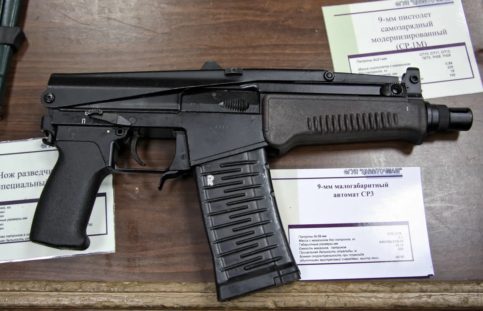 A compact black submachine-gun-like carbine with a folded top-mounted stock and a curved magazine, lying on a wooden table beside two Russian-language exhibition placards.