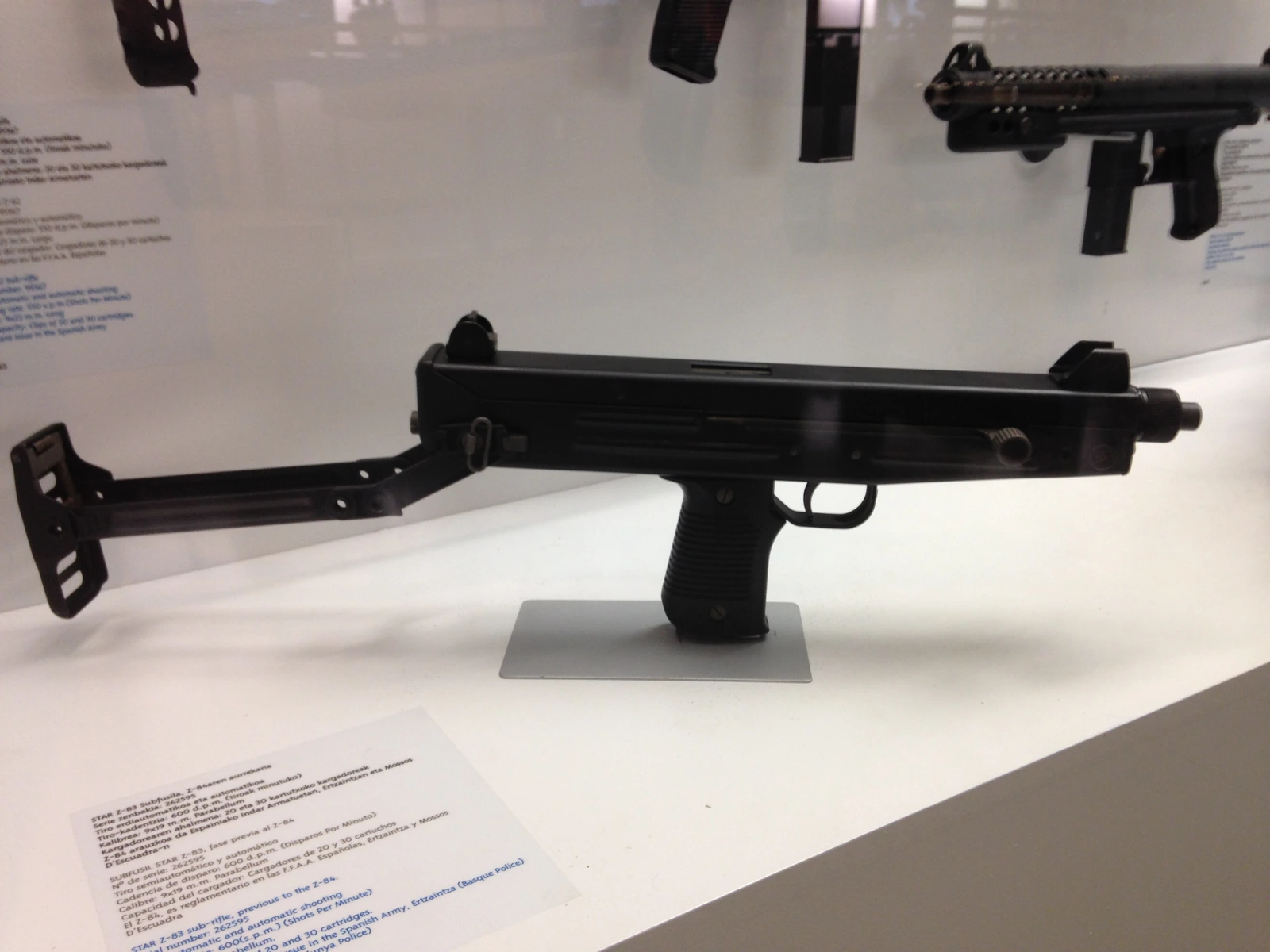 A black submachine gun with a folded stock and a straight box magazine, standing upright on a museum display stand