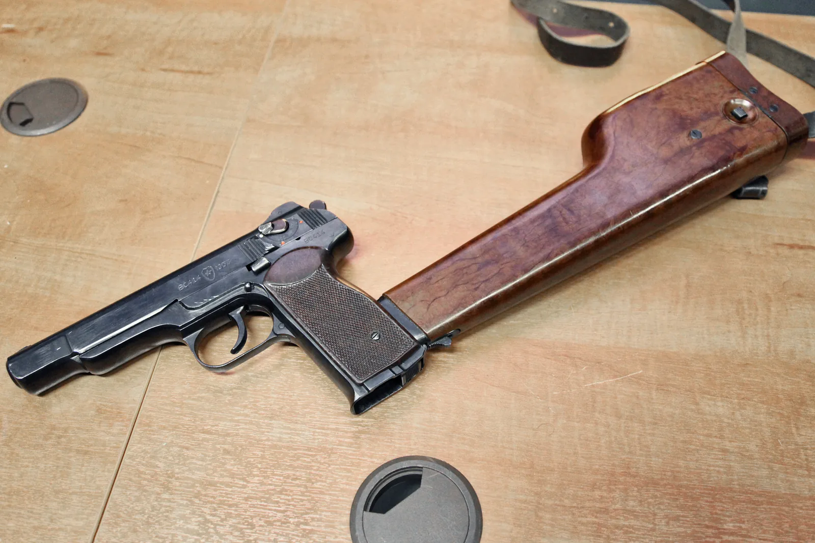 A blued Soviet pistol lying on a plywood bench, seen from the left at an angle. It is unusually long-slided for a pistol, with a chequered brown grip, a slide-mounted selector lever and a large box magazine standing proud of the butt. Attached to the rear of the grip is a hollow wooden holster used as a shoulder stock, longer than the pistol itself.