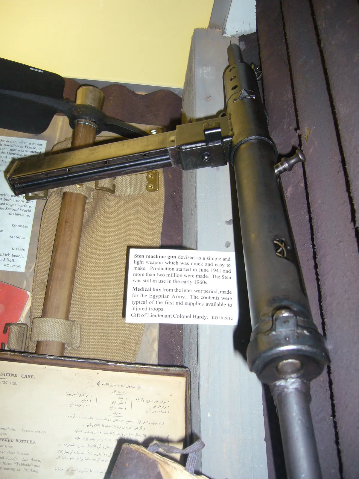 A dark-finished Sten Mk II submachine gun seen from above and slightly to the rear, showing its plain tubular receiver, side-mounted magazine housing and skeletal wire shoulder stock