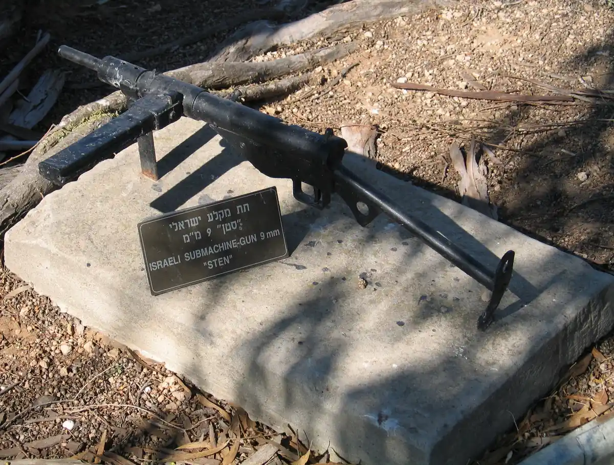 A black Sten Mk II submachine gun with a T-shaped wire stock, propped up outdoors on a stony, dirt-covered ground beside a small plaque