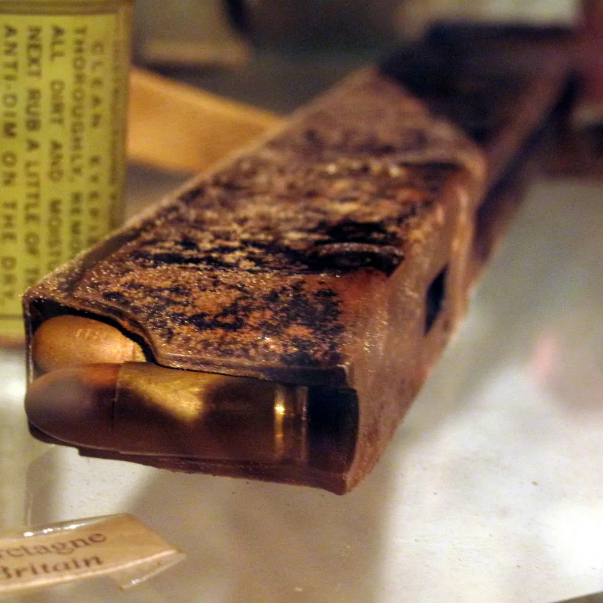 Close-up of a long, straight, double-column steel box magazine for a Sten submachine gun, standing on end with a row of cartridges visible through its feed lips