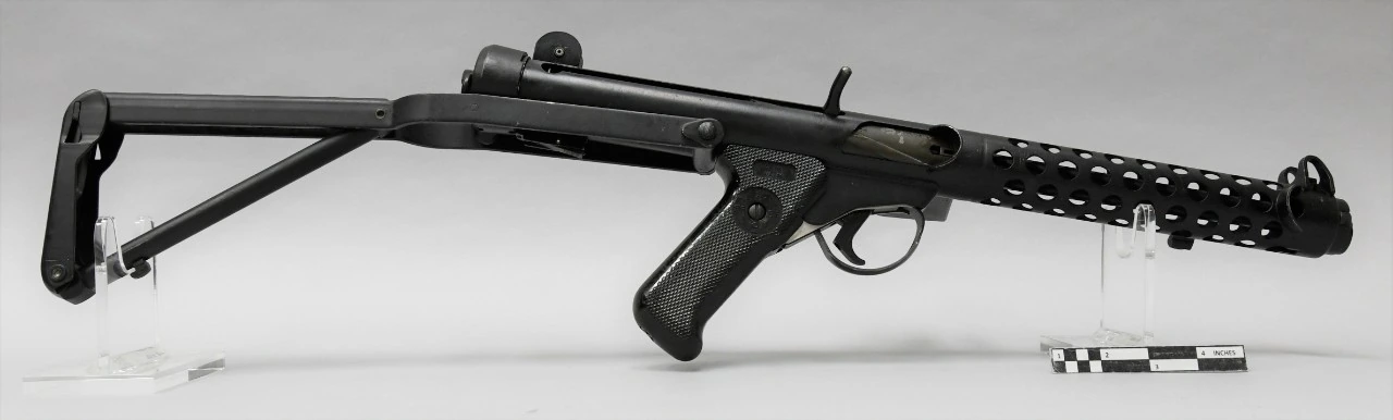 A Sterling submachine gun on museum stands against a grey background, seen from the left with the stock folded under, showing a perforated tubular receiver and a black plastic pistol grip