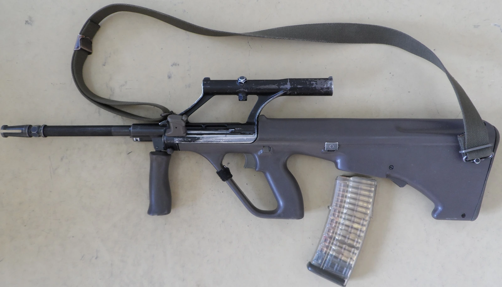 A Steyr AUG rifle lying on a pale surface with a green sling, seen from the left: a green polymer bullpup body, a carrying handle containing an optical sight, a folding forward grip, and a translucent magazine lying beside it
