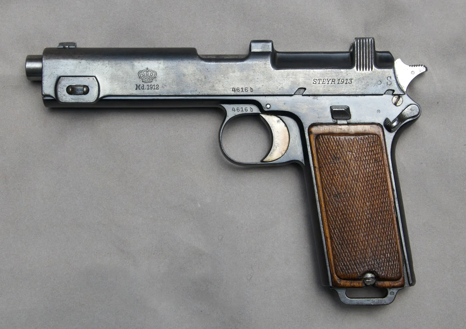A blued semi-automatic pistol on a grey ground, seen from the left. The slide is short and sits in a cut-out at the rear of a long one-piece frame; an external hammer stands at the back. The grip carries chequered wooden panels. Markings on the frame read Md.1912 under a crown, and STEYR 1913 on the slide.