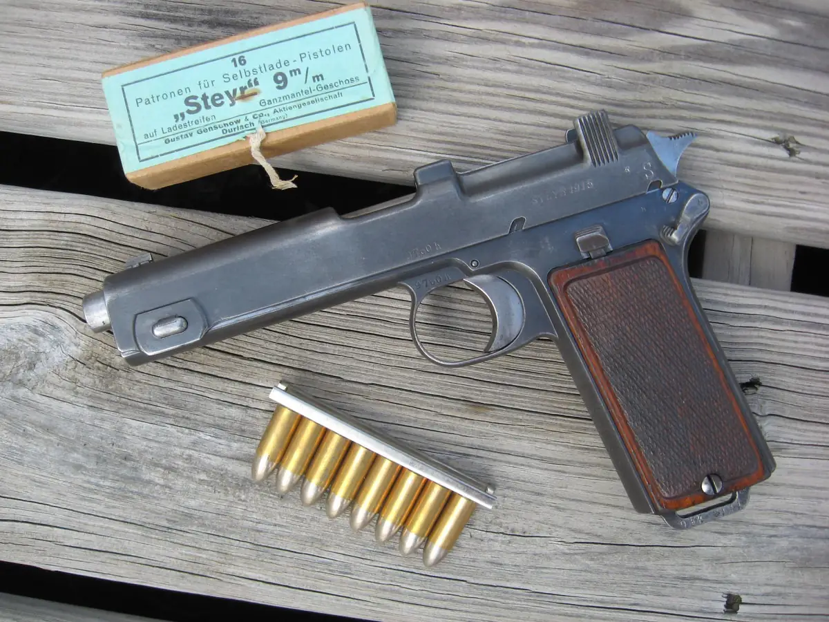 A Steyr M1912 pistol lying on weathered grey planks, seen from the right, beside a green cardboard ammunition packet in German and a metal charger clip holding eight brass cartridges with round-nosed bullets.