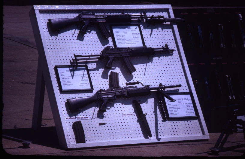 Several rifles and a belt-fed light machine gun of the same weapon family displayed together on a pegboard, alongside printed information cards.