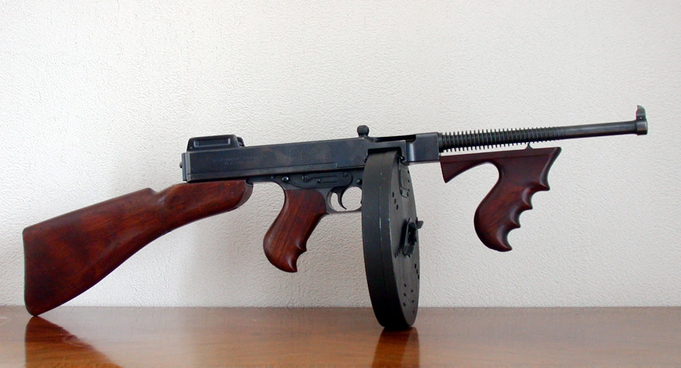 A Thompson submachine gun standing on a wooden table, with a walnut butt, two wooden grips, a finned barrel and a large circular drum magazine below the receiver