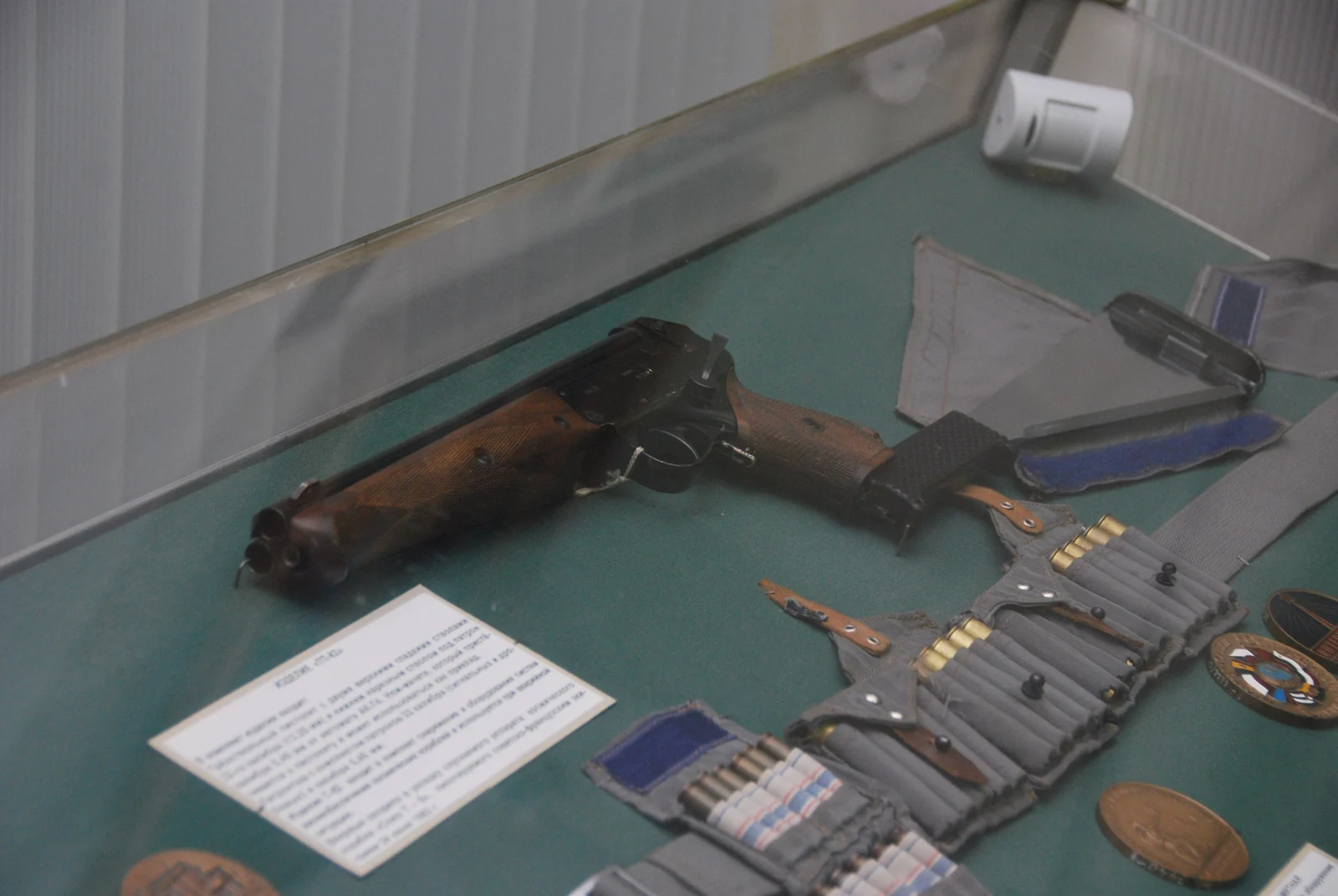 A dark, blued triple-barrelled pistol with a wooden grip and no buttstock attached, displayed in a museum case beside an ammunition bandolier and its canvas machete sheath.