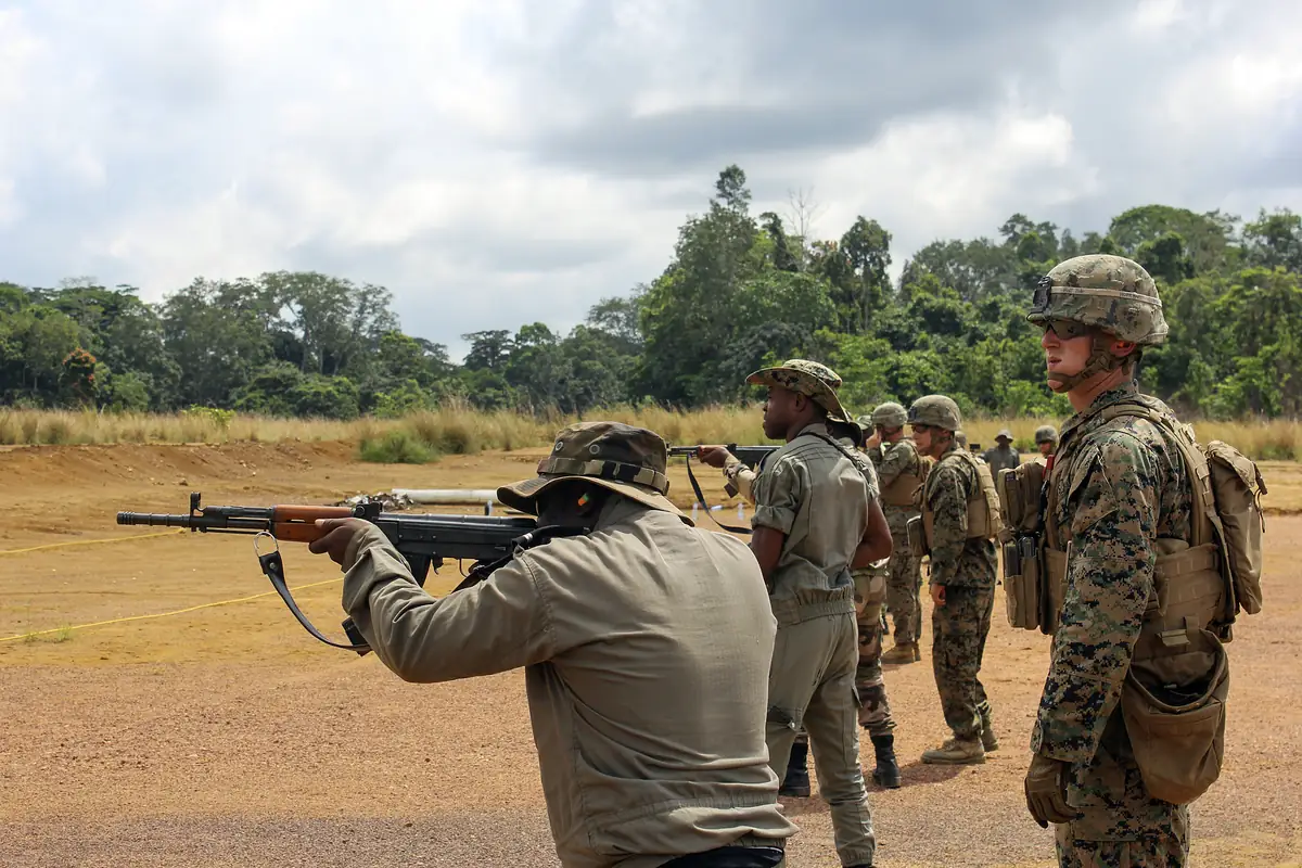 A soldier in tan uniform fires a rifle at a paper target on a shooting range while a US Marine observes beside him.