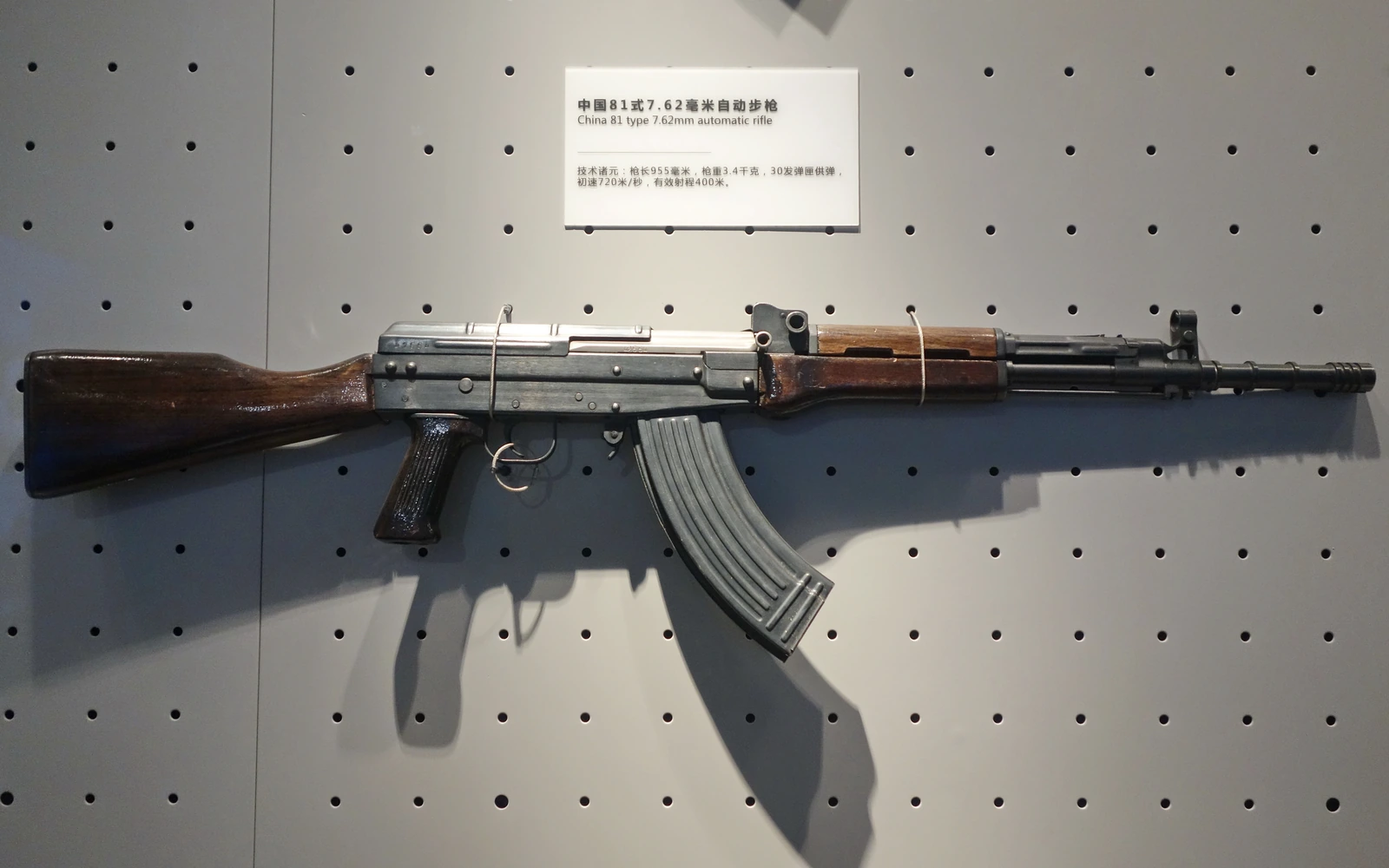A rifle with a wooden handguard, a curved 30-round magazine and a folding metal wire stock, mounted on a museum display panel.