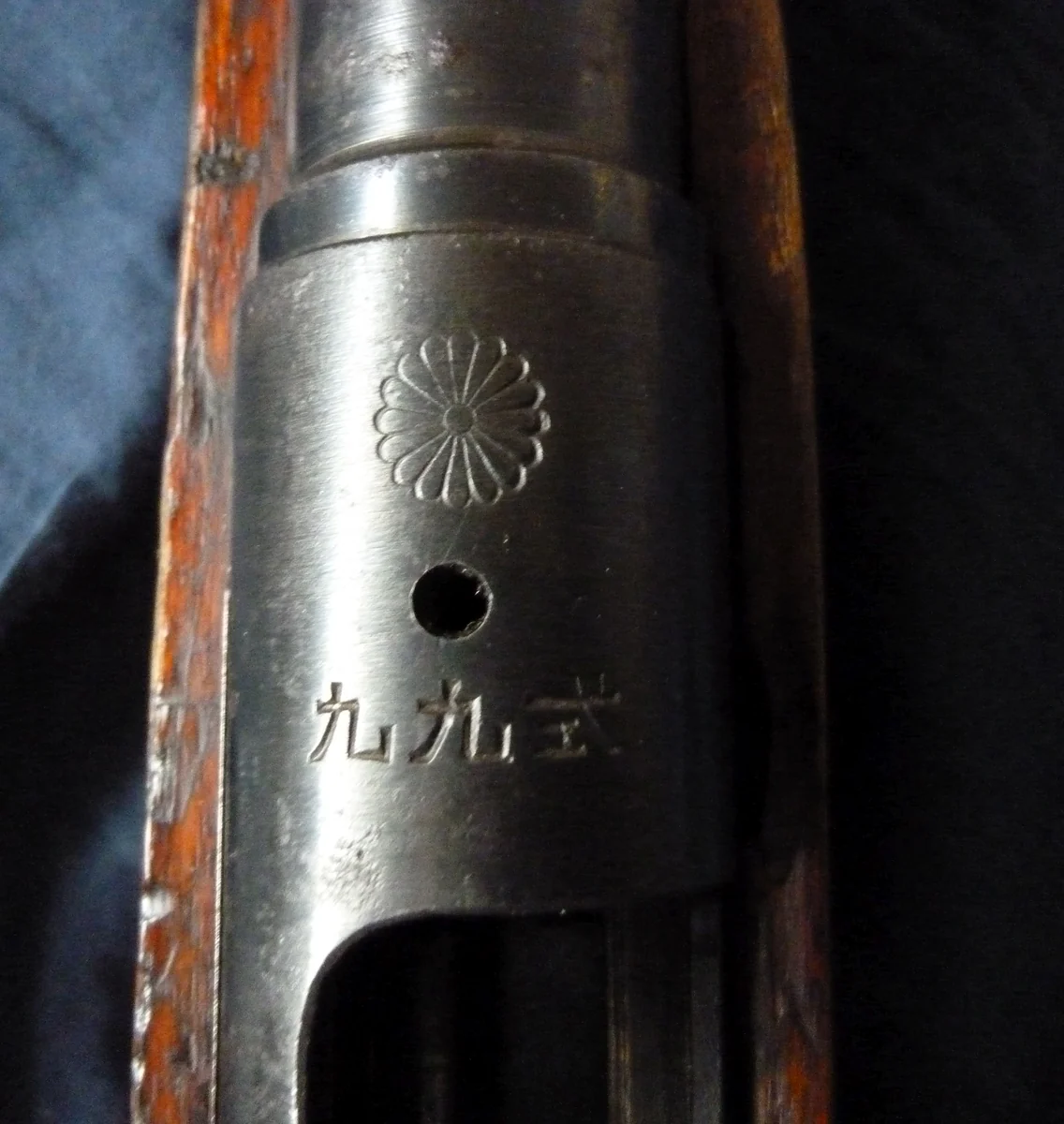 A close view looking down on the front of a rifle receiver, stamped with a sixteen-petal chrysanthemum above a single round vent hole and three Japanese characters.