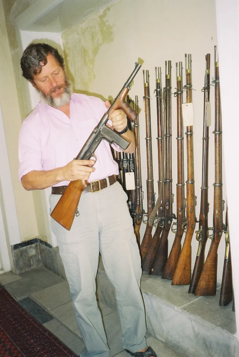 A man holding an M42 submachine gun beside a rack of older bolt-action rifles