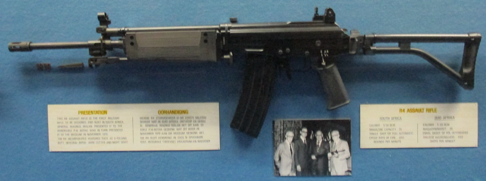 An early-production rifle with a folding wire stock, a curved box magazine and a ribbed handguard, displayed in a museum case with a period photograph beside it.