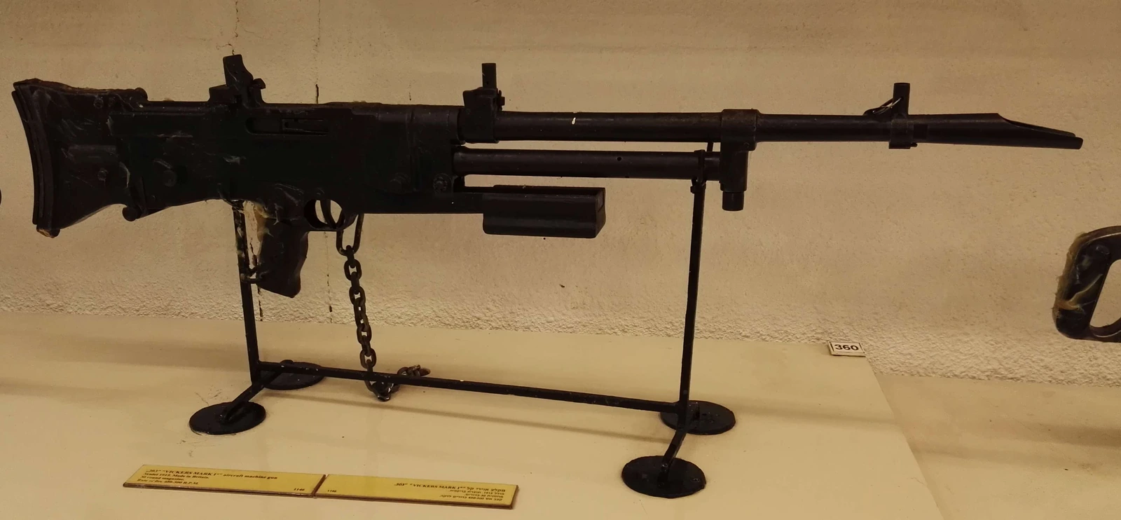 A black machine gun with a distinctive perforated barrel jacket, a spade grip and a large pan-shaped magazine on top, mounted on a museum stand