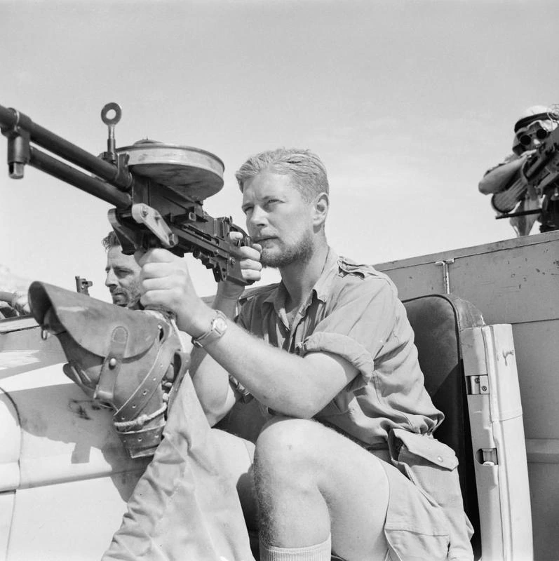 A soldier sits behind a Vickers K machine gun mounted on a truck, with a second gun visible mounted beside him, in a desert setting