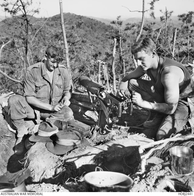 Two soldiers crouched outdoors cleaning a Vickers machine gun, its barrel jacket and ammunition belt visible on the ground beside them