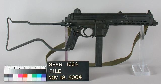 A gray-green metal submachine gun with a folded wire stock, a curved magazine, and a museum inventory placard beneath it