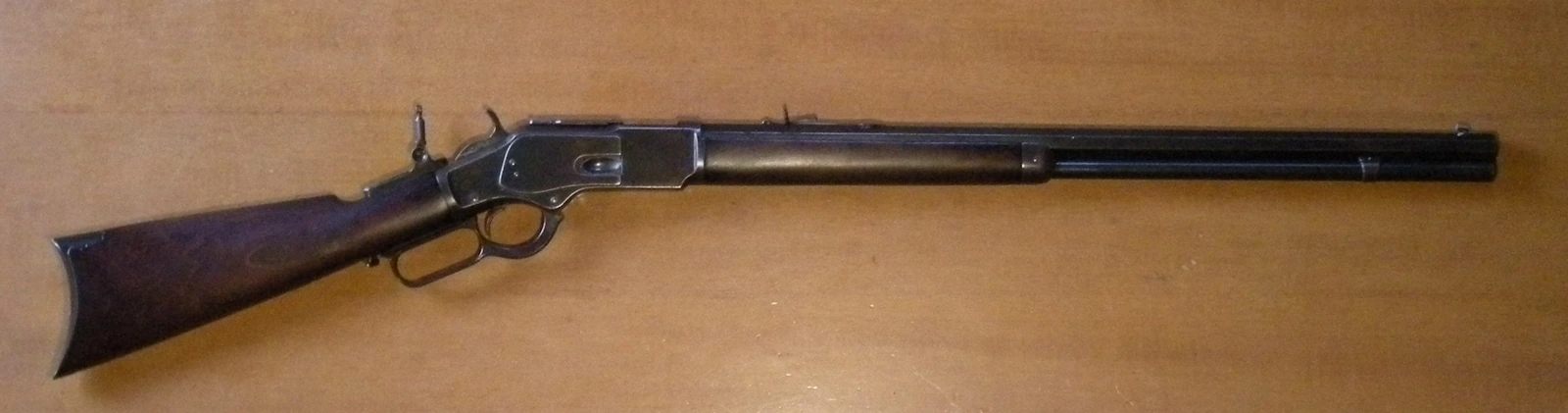 A Winchester Model 1873 rifle lying on a wooden surface, seen from the left, with a walnut stock, a colour-cased receiver, an octagonal barrel and a loading lever below the trigger