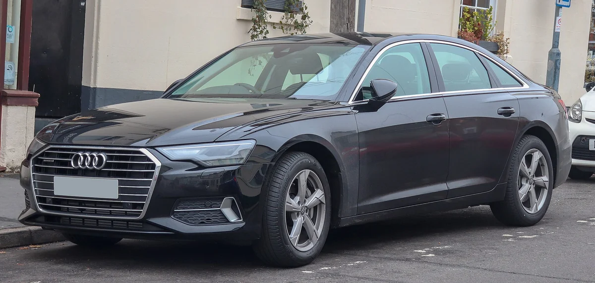 Black Audi A6 C8 saloon photographed from the front three-quarter angle on a street, with a wide chrome-edged single-frame grille and slim LED headlights