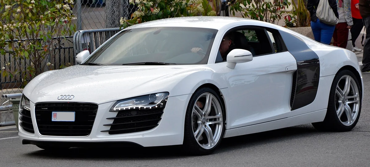 White first-generation Audi R8 coupe photographed from the front three-quarter angle on a street, with a low wide grille bearing the four-rings badge and LED daytime running lights along the intake