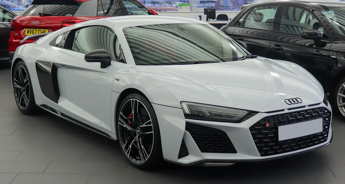 White facelifted second-generation Audi R8 coupe photographed from the front three-quarter angle in a showroom, with an angular hexagonal grille bearing a V10 badge on the door sill