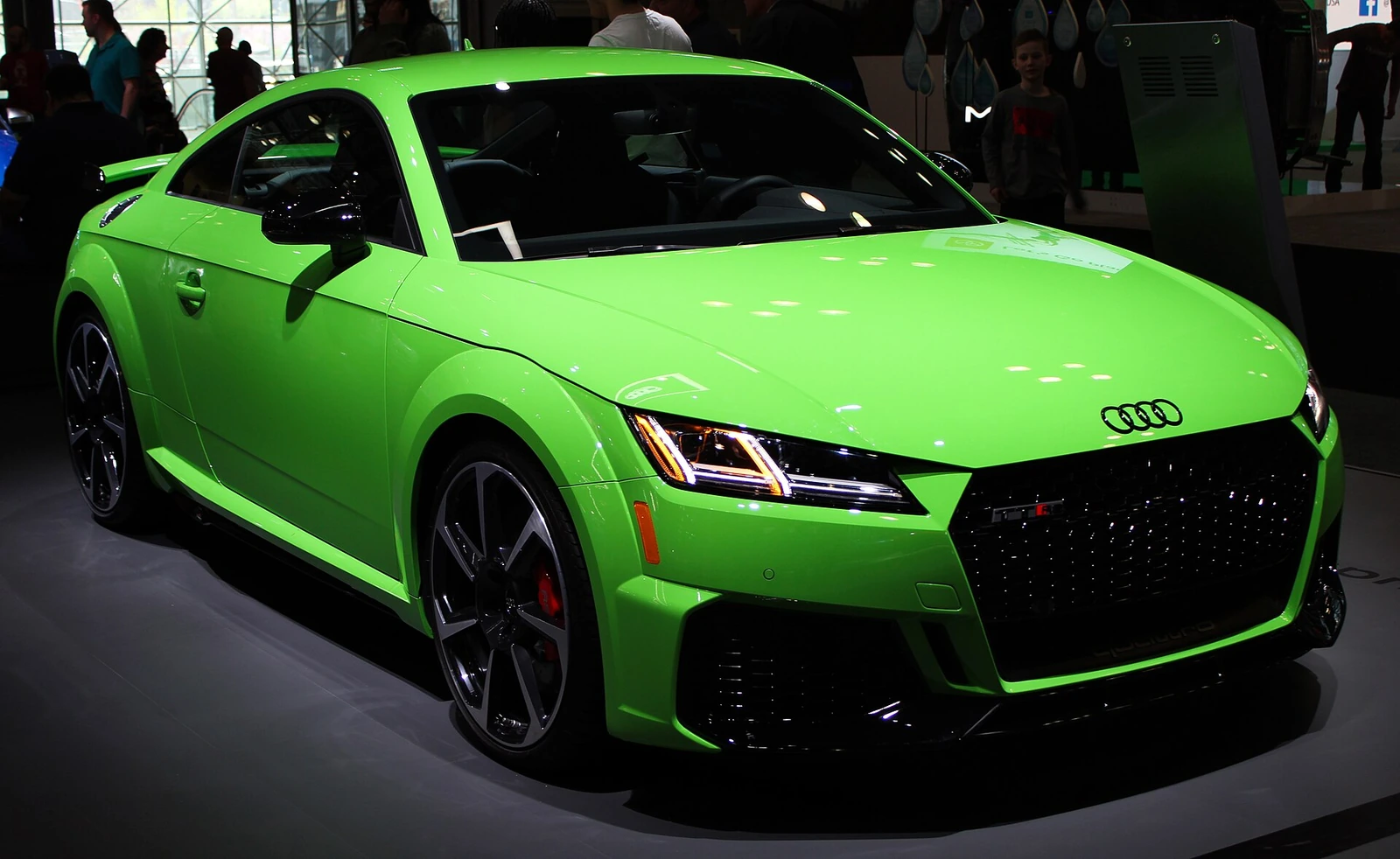 Green Audi TT RS with technical specifications displayed alongside it at a motor show, photographed from the front three-quarter angle