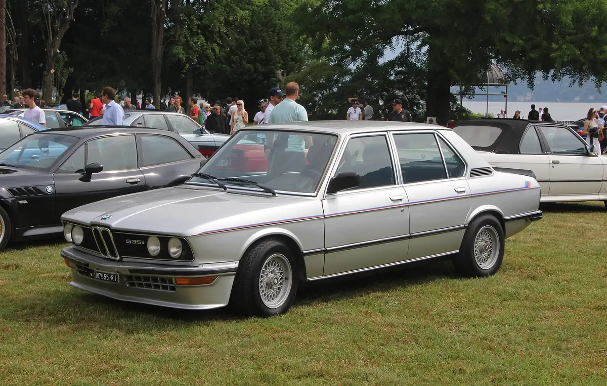 Silver BMW E12 M535i saloon with a tricolour side stripe, photographed from the front three-quarter angle at a car show