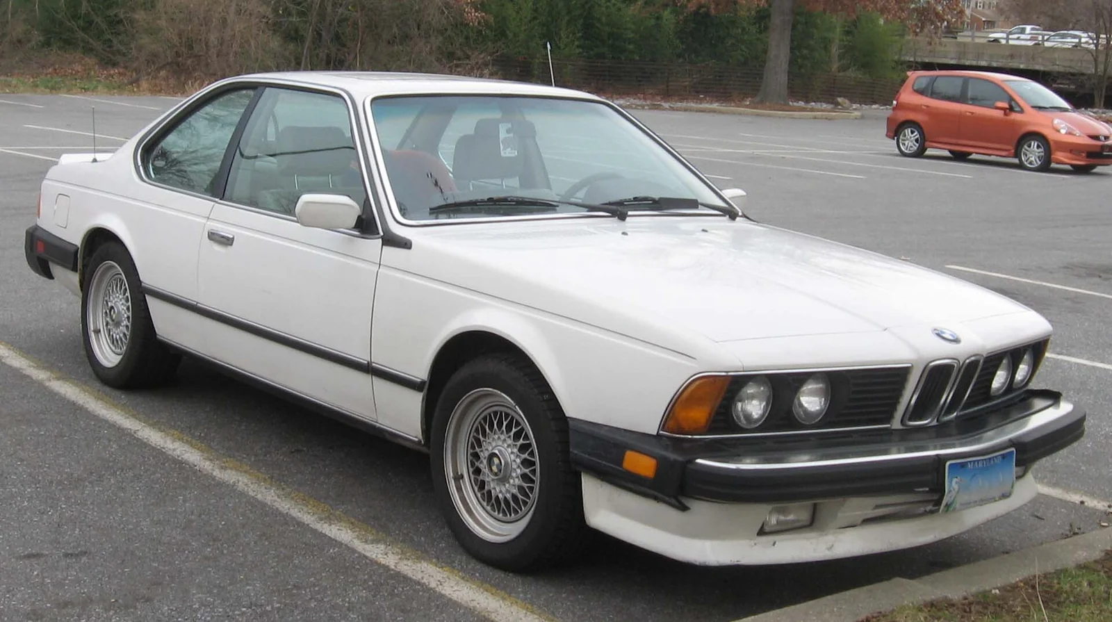 White BMW 6 Series E24 coupé photographed from the front three-quarter angle, showing the shark-nose front and the large impact bumpers of a US-market car