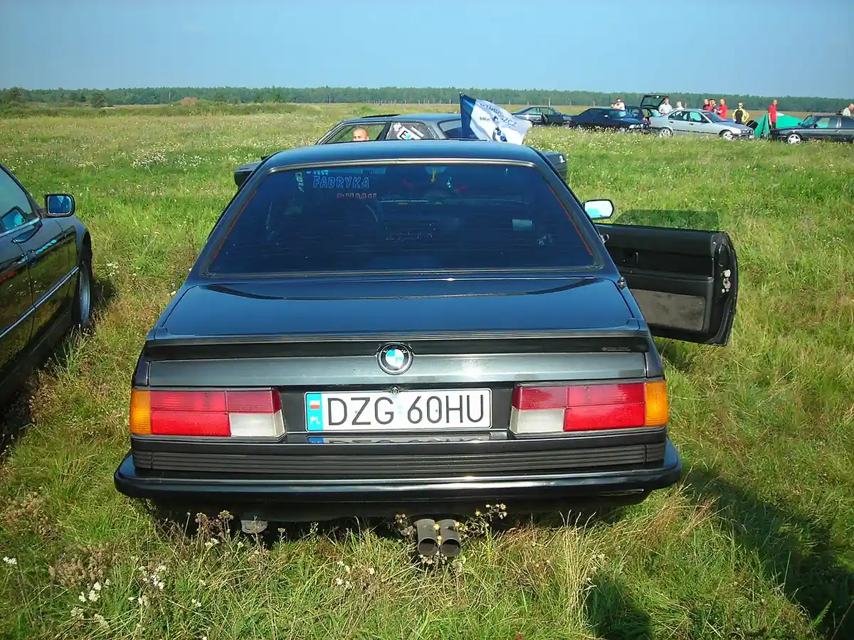 Rear view of a dark grey BMW 6 Series E24 in a grass field at a car meet, with the driver's door open