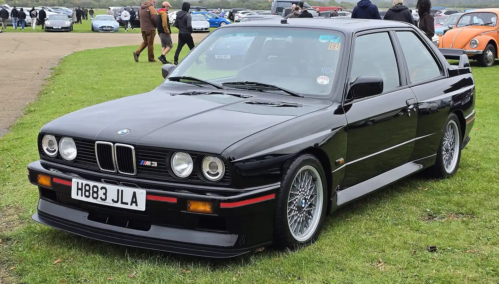 Black BMW E30 M3 coupe photographed from the front three-quarter angle on grass at a car show