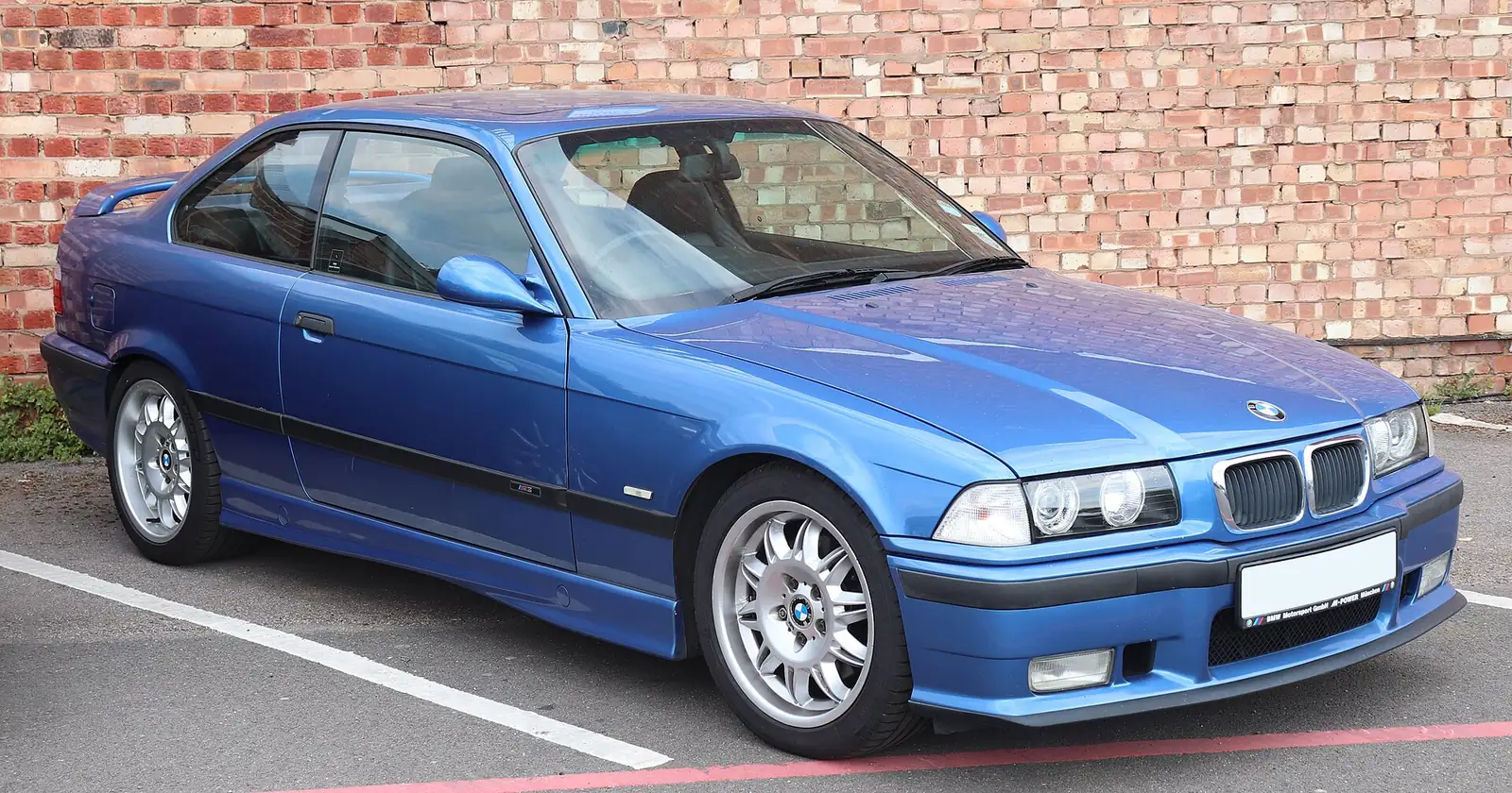 Blue BMW E36 M3 coupe photographed from the front three-quarter angle, parked against a brick wall