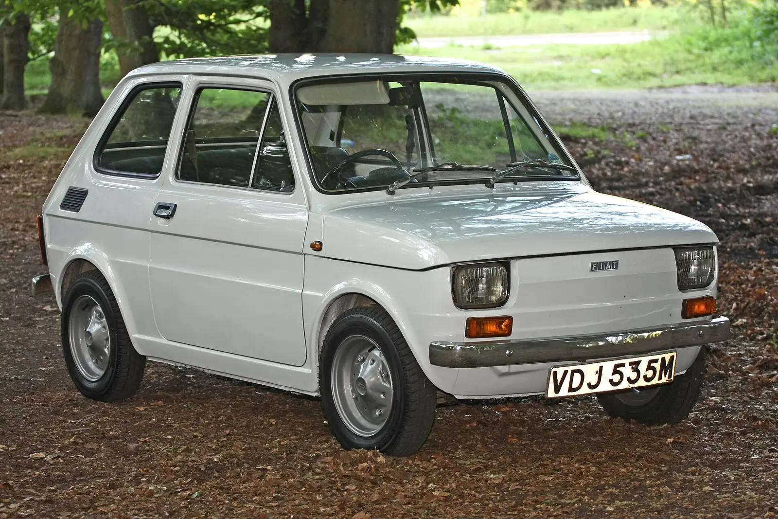 White Fiat 126 photographed from the front three-quarter angle, showing its boxy two-door body and round headlights