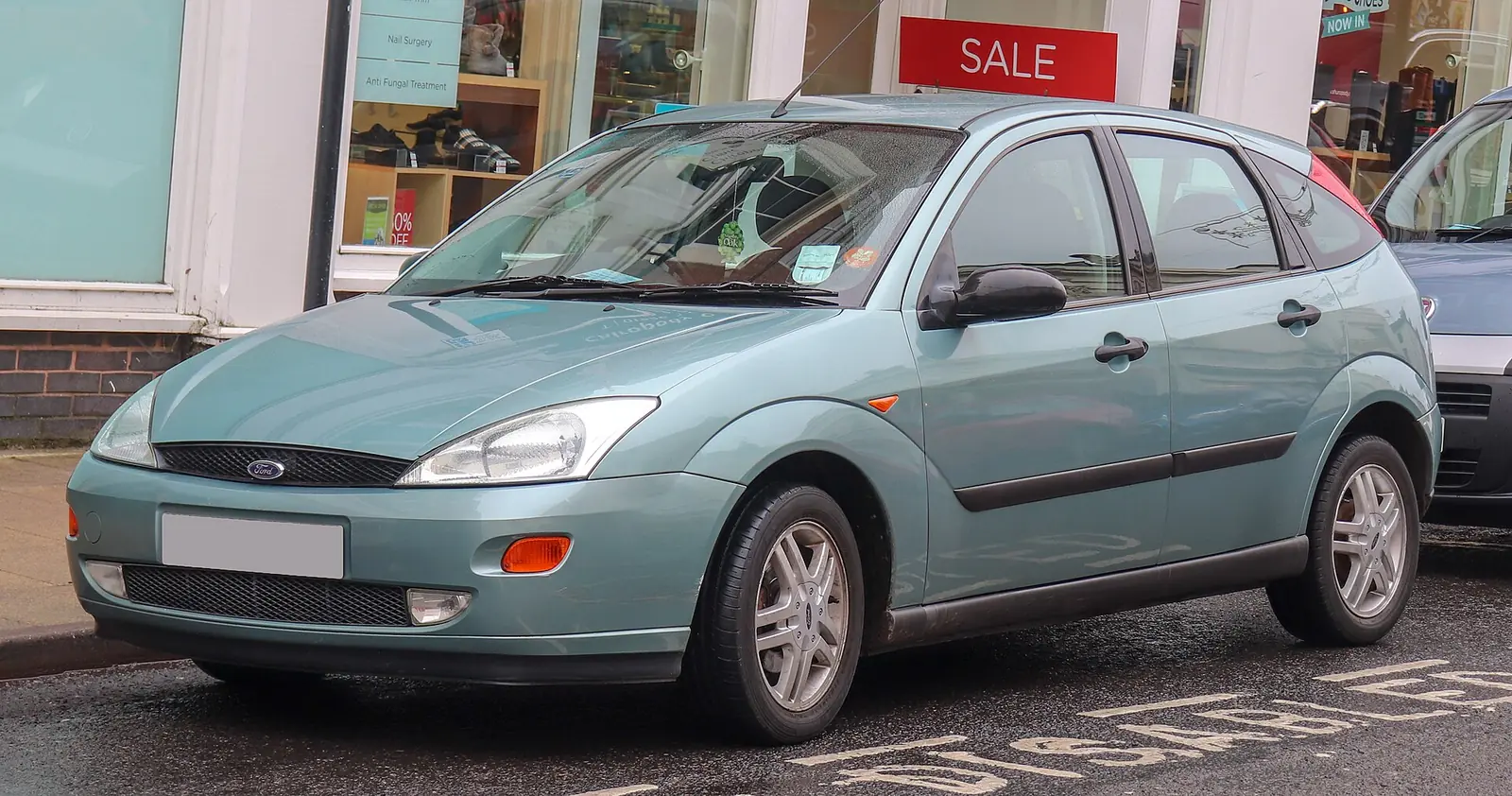 Pale green first-generation Ford Focus hatchback photographed from the front three-quarter angle outside shops