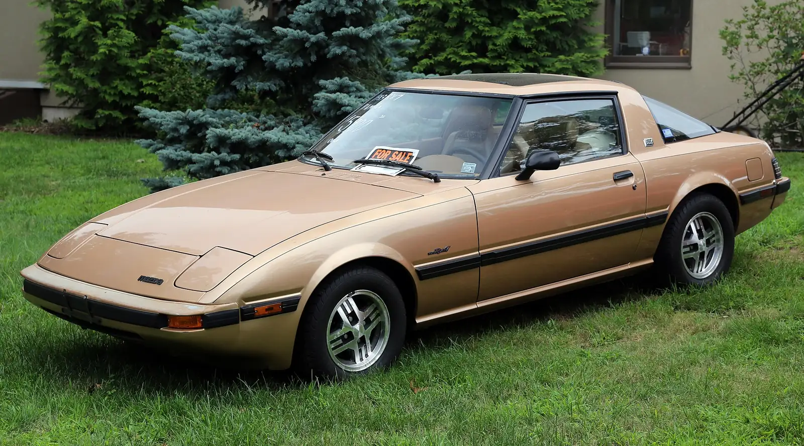 Gold first-generation Mazda RX-7 GSL photographed from the front three-quarter angle, showing its pop-up headlight covers and low, wedge-shaped nose