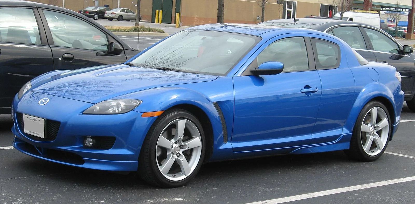 Blue pre-facelift Mazda RX-8 photographed from the front three-quarter angle in a car park, showing its four-door quad-coupe silhouette with no visible B-pillar