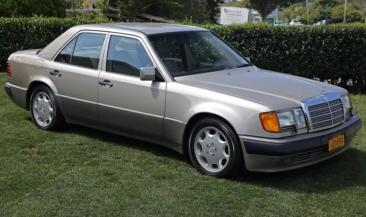 Gold-coloured Mercedes-Benz 500E saloon photographed from the front three-quarter angle on grass, with wide fender flares and multi-spoke alloy wheels