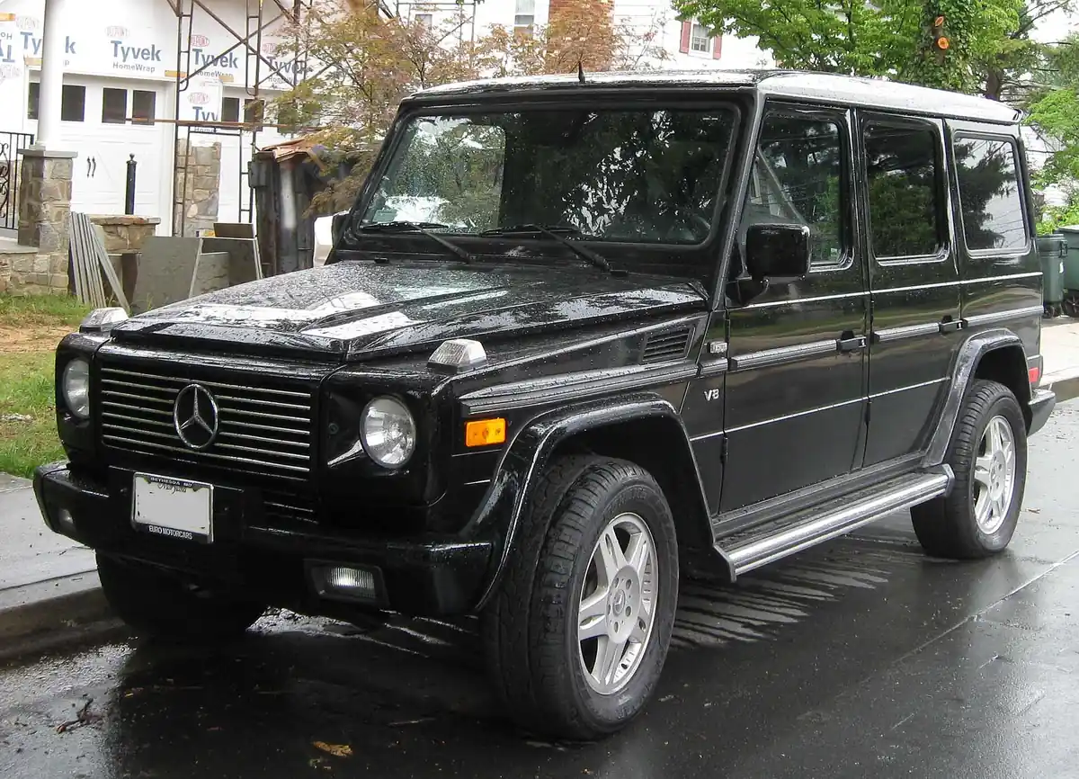 Black first-generation Mercedes-Benz G-Class SUV photographed from the front three-quarter angle on a wet street, with a boxy body and a large chrome grille bearing a central star