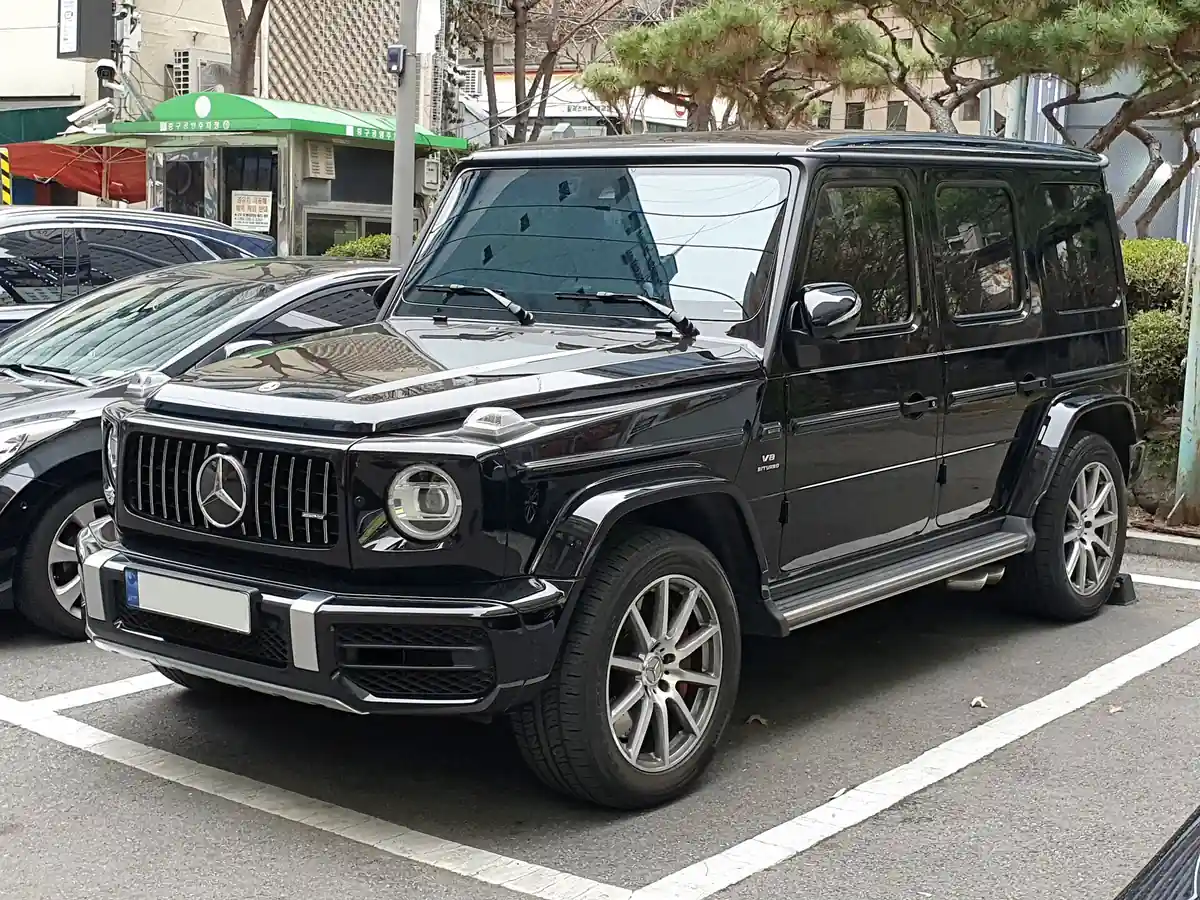 Black second-generation Mercedes-AMG G 63 photographed from the front three-quarter angle in a car park, with a wide chrome-slat grille, round headlamps and prominent side exhaust pipe
