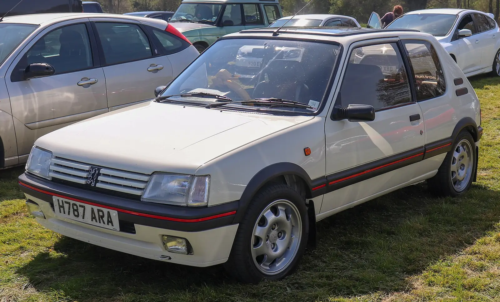 Cream Peugeot 205 GTI 1.9 hatchback photographed from the front three-quarter angle, parked on grass among other cars